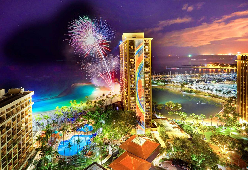 Hilton Hawaiian Village and its iconic Rainbow Tower in view at night with fireworks offshore.